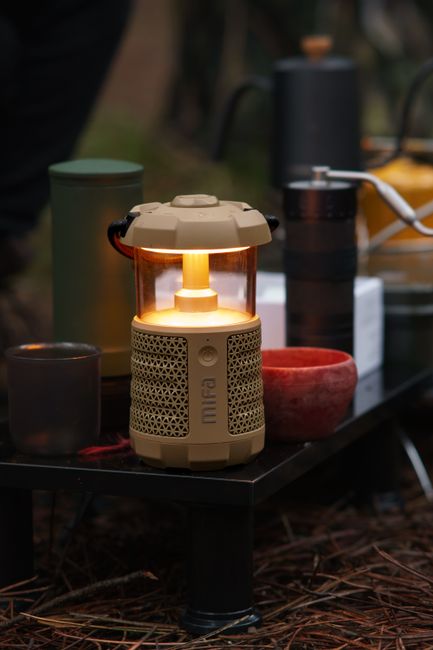 A close-up of a glowing portable electric lantern on a camping table with cups and equipment outdoors.