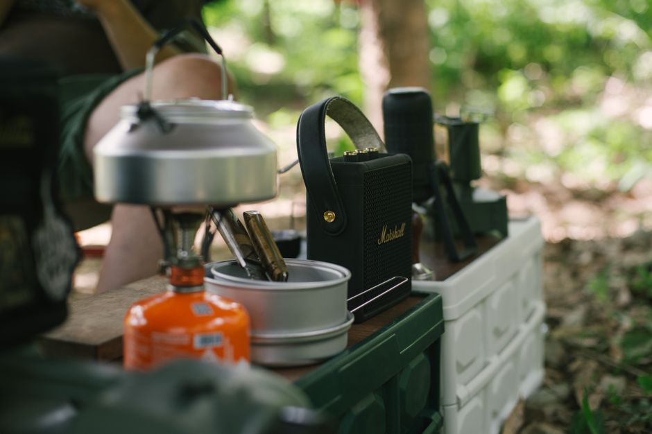 Outdoor camping gear including a gas stove and kettle on a table in a forest setting.