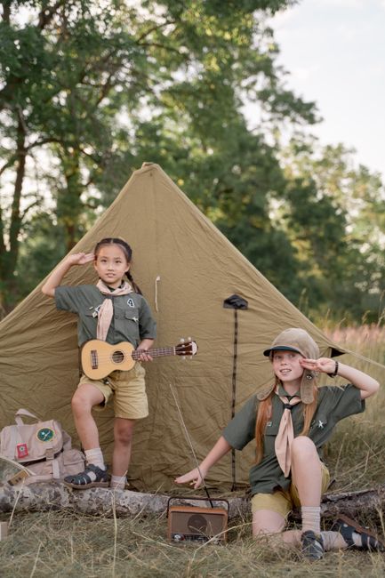 Two girls in scout uniforms salute in front of a tent, one holding a ukulele, at a scouting camp.