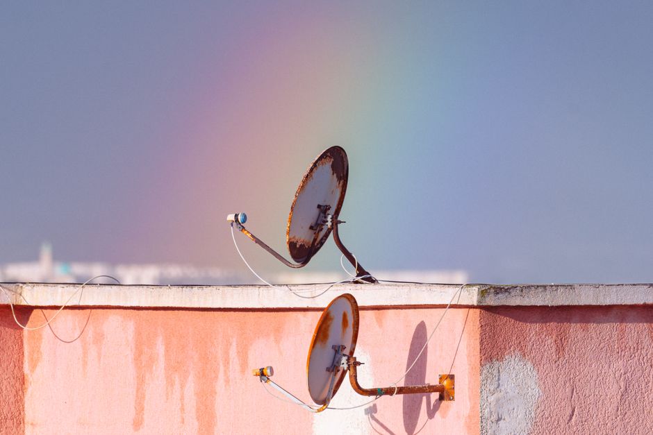 Two rusty satellite dishes on a rooftop against a vibrant rainbow in Tunisia.